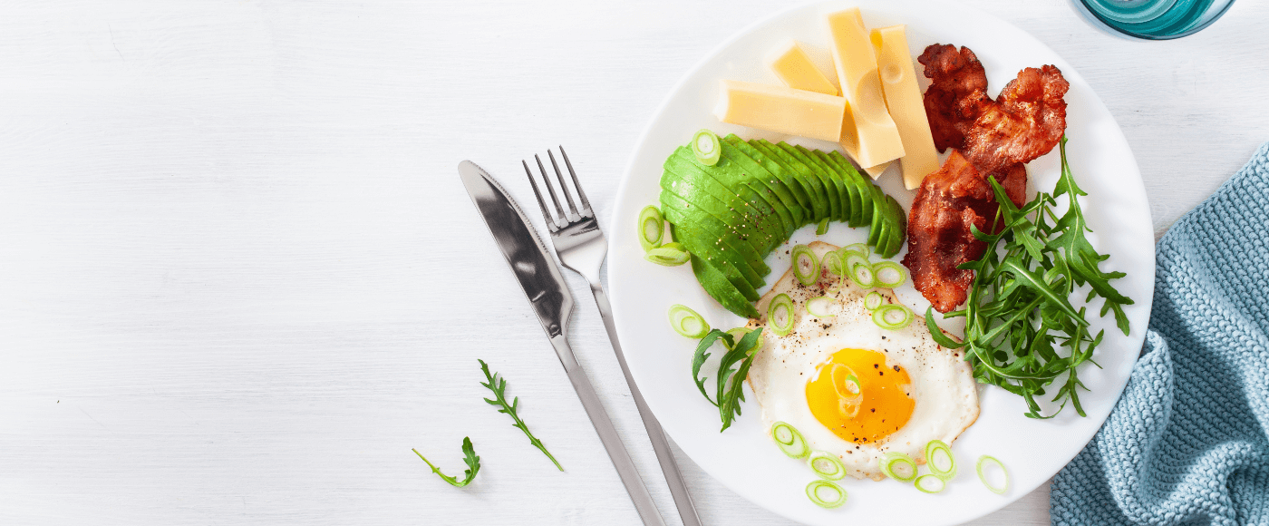a breakfast plate with avocado slices, fried egg, bacon, cheese, arugula & green onions set on a white table