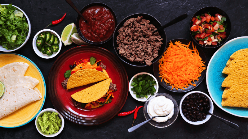 taco bar table scene with a variety of ingredients such as tortillas, sour cream, minced beef, salsa etc. on dark slate background