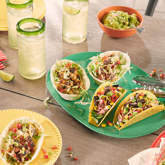 Various types of tacos placed in a dish along with glasses filled with juice placed on a wooden table.