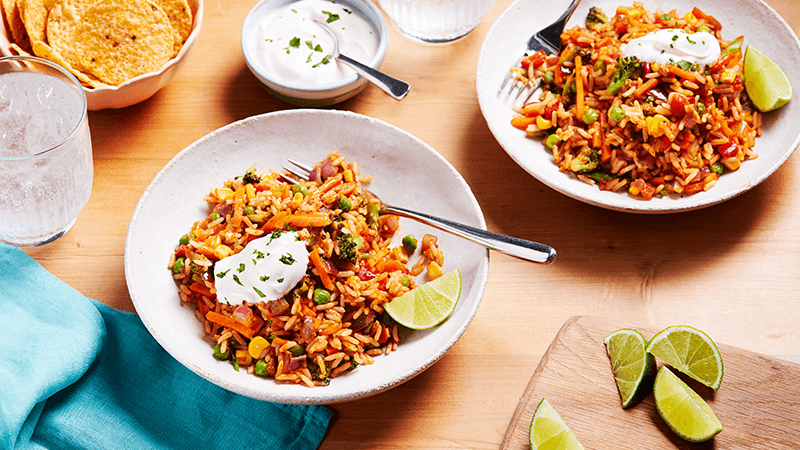 Two bowls of Veggie Mexican Rice Bowls with a dollop of soured cream on top, surrounded by chips, soured cream and lime wedges